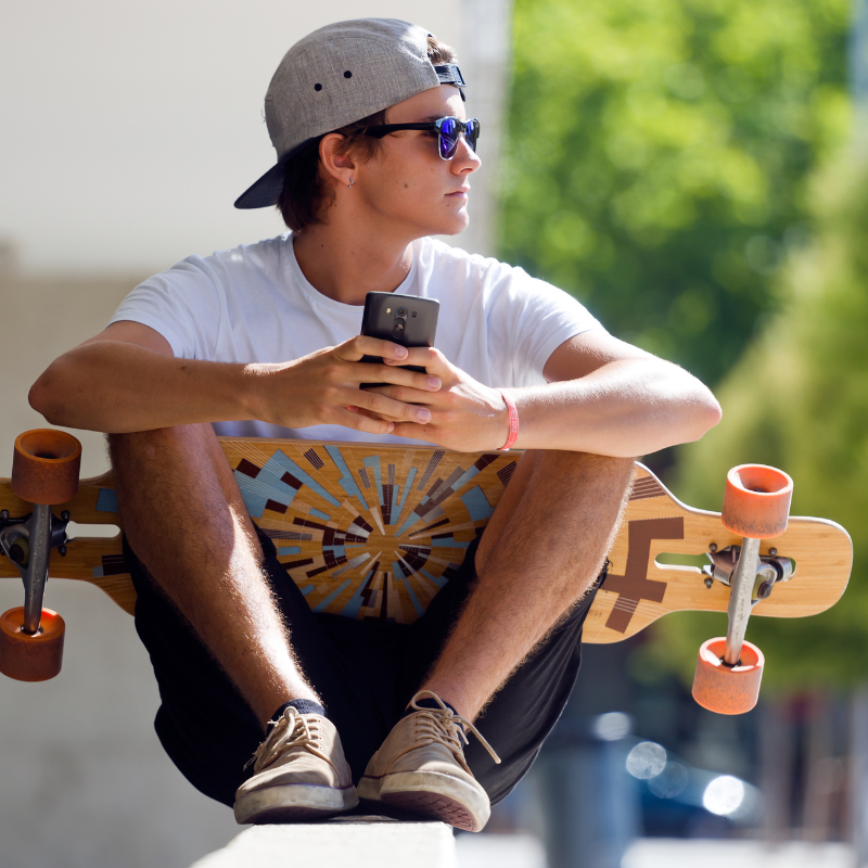 A trendy young guy sits with a longboard skateboard and holds his phone in his hands.