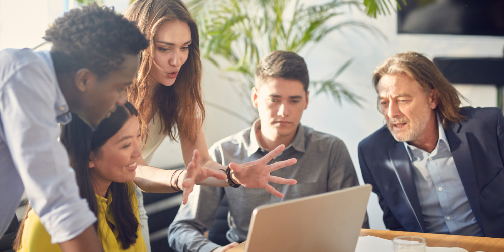 A group of five professionals are gathered around a computer and having a lively discussion while looking at the screen