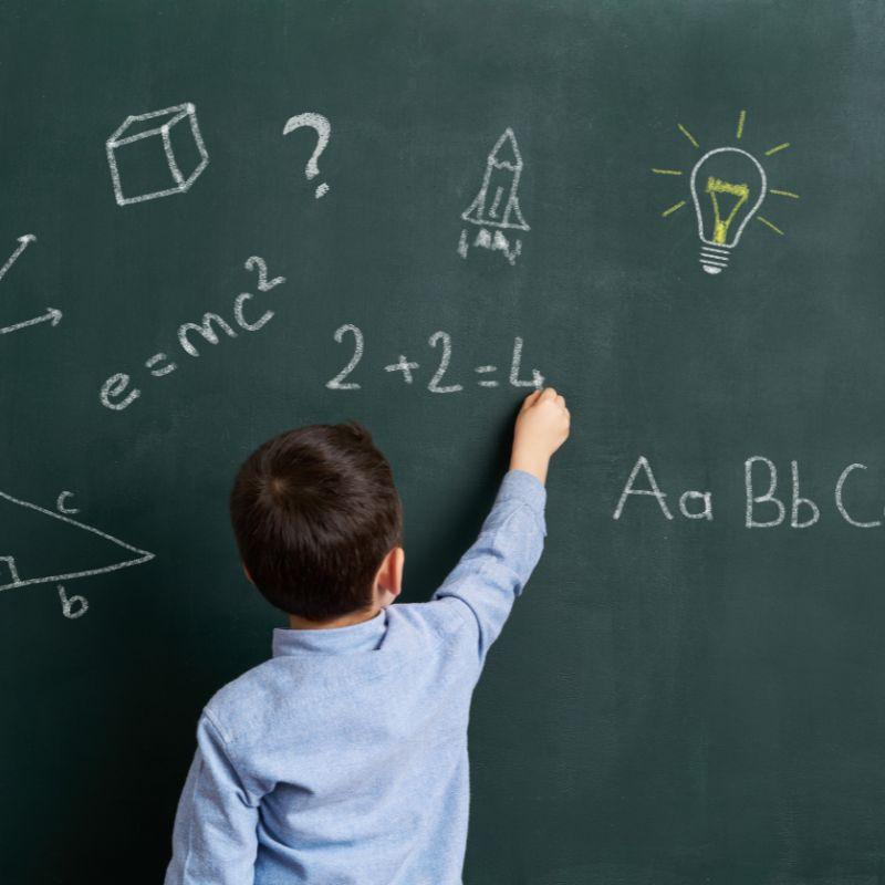 Niño de espaldas escribiendo en una pizarra, concentrado en resolver un problema matemático en clase.