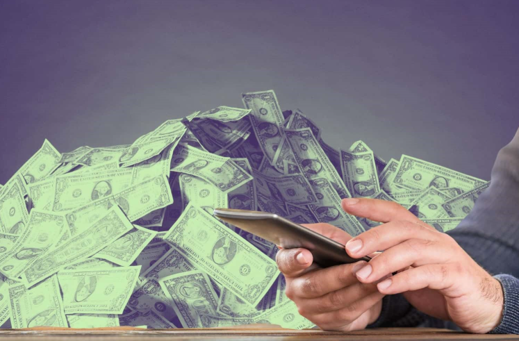 A person typing on a cell phone and a stack of cash in the background 