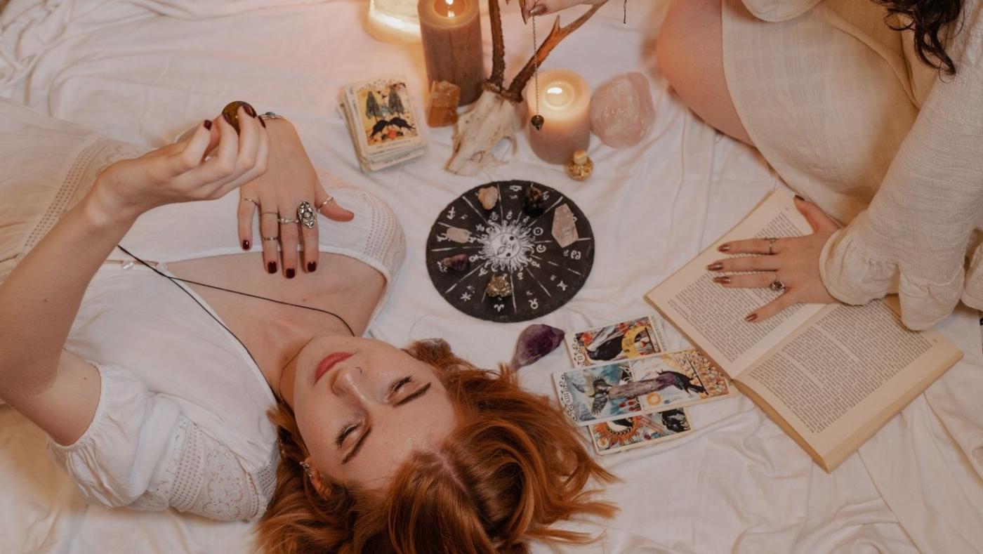 Imagen de dos mujeres jovenes sobre una manta con un tablero de astrología, un libro, un oráculo y velas. 