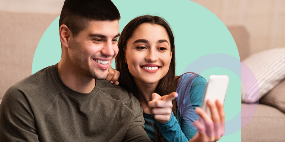 A man and a woman are watching a livestream together on their phone. The woman points to the phone, and they're both smiling.