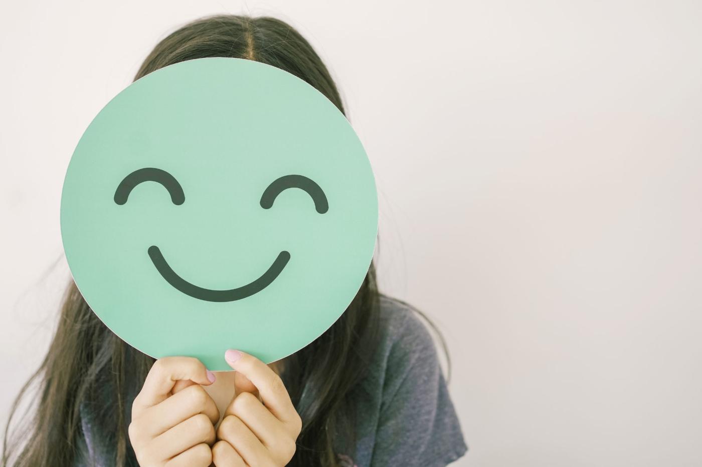 Imagen de una persona tapando su cara con un círculo de cartón con una carita sonriente, representando la problemática de la salud mental.