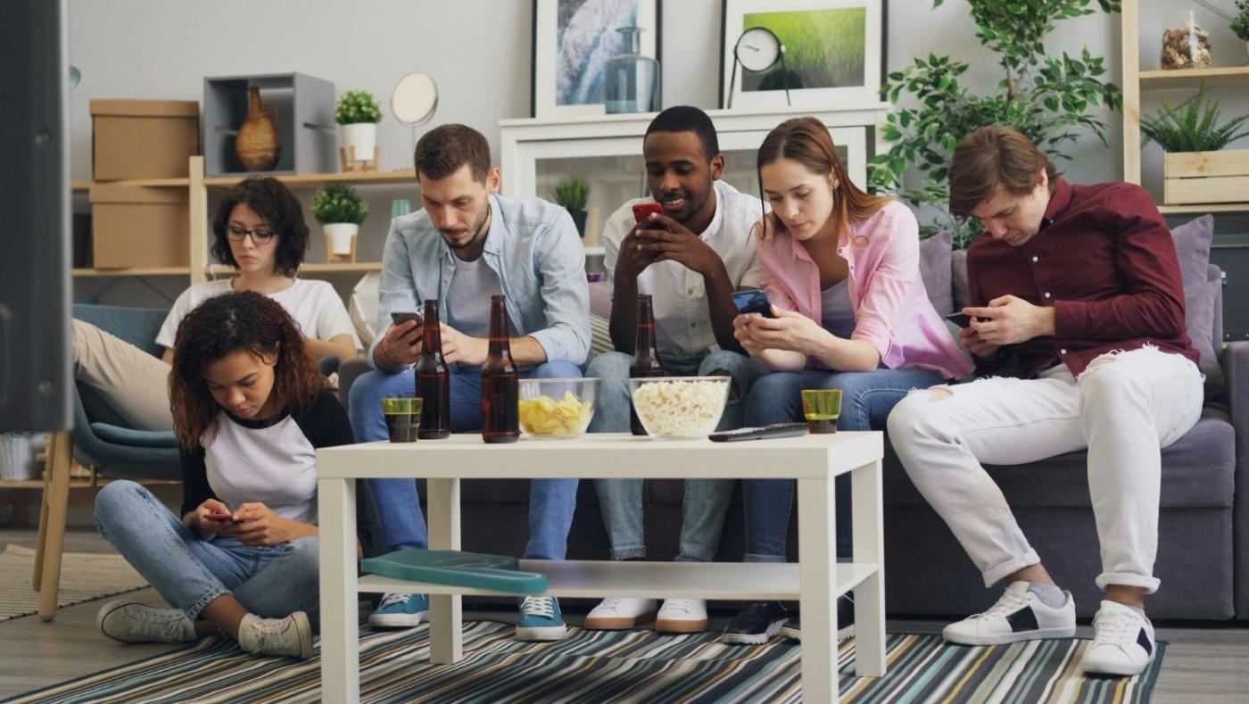 Grupo de amigos reunidos en una mesa mientras varias personas miran sus teléfonos celulares en lugar de conversar
