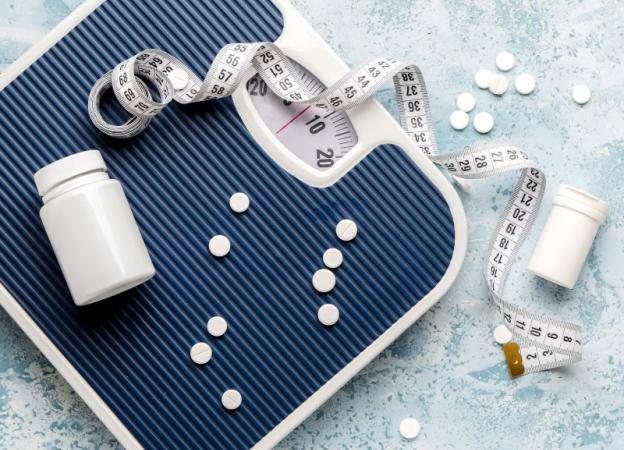 A blue and white bathroom scale surrounded by a tape measurer, two pill bottles, and some round white pills. Weight loss theme.