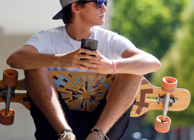 A trendy young guy sits with a longboard skateboard and holds his phone in his hands.