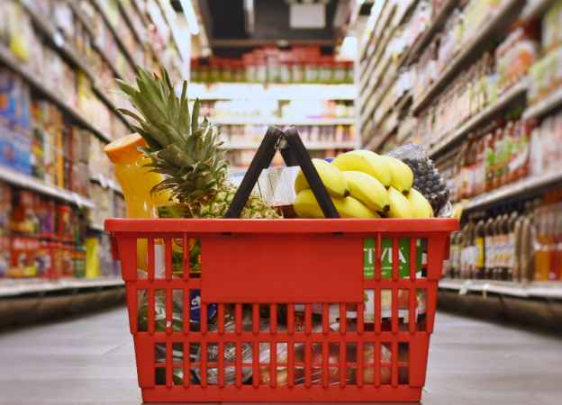 A red shopping basket filled with groceries sits on the floor in the aisle of a grocery store