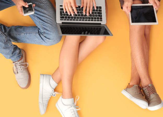 Three people sitting on a yellow background. One is using a phone, one a laptop, and one a tablet.